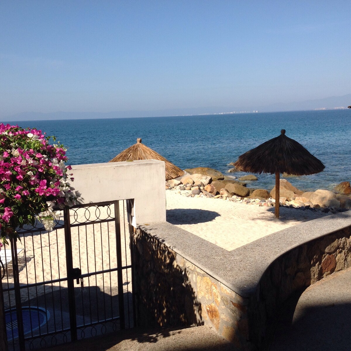 A serene beachfront view is presented, featuring sandy shores leading to calm waters. Thatched umbrellas offer shaded spots on the sand, while a colorful flowering plant frames the entrance, adding a touch of vibrancy to the coastal scene.