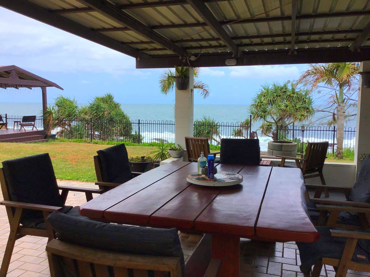 An outdoor dining area features a large wooden table surrounded by chairs, all under a sheltered roof. The ocean view stretches beyond the grassy yard, with palm trees framing the scene. Nearby, a shaded lounge space is visible, providing additional seating.
