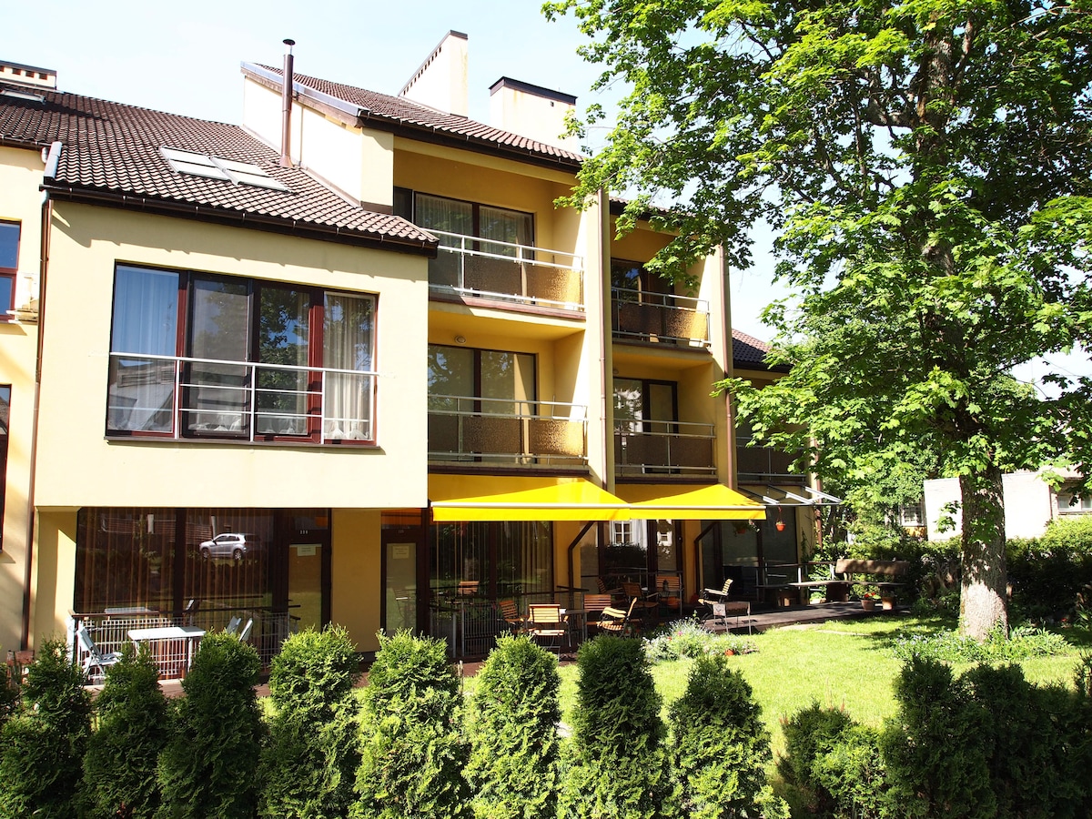 The exterior of the building features a modern design with multiple balconies, each adorned with yellow awnings. A well-maintained green lawn is framed by neatly trimmed shrubs, providing a pleasant outdoor space. Large windows allow ample natural light into the apartments.