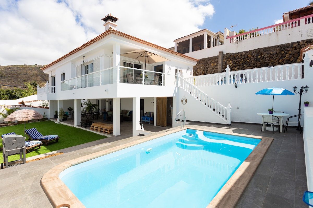 The villa features a private outdoor space with a turquoise swimming pool surrounded by gray tile. Sun loungers and a shaded area with an umbrella are visible. The two-story structure overlooks the pool, with a spacious terrace accessible from the upper level.