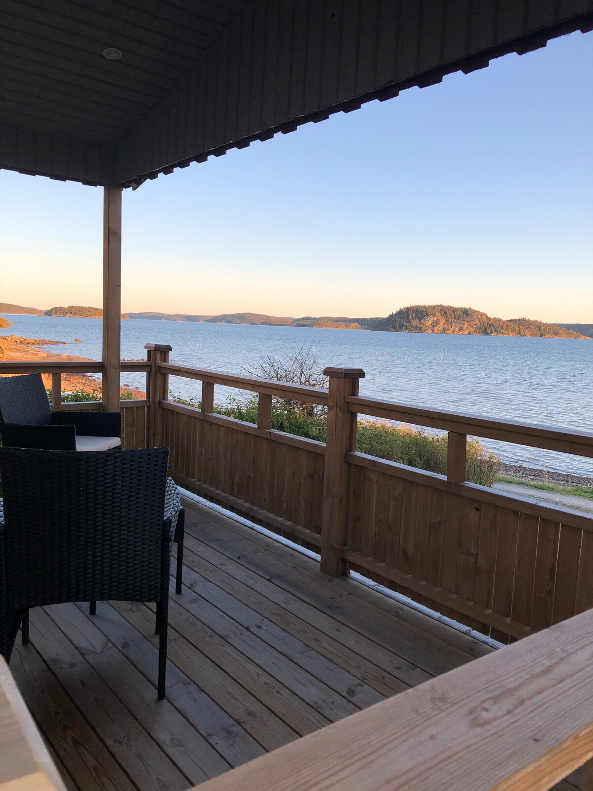 A wooden deck offers a clear view of the water and the distant landscape. Black wicker chairs surround a table, creating a relaxed outdoor seating area. The soft colors of the sunset illuminate the scene, enhancing the natural beauty of the surroundings.