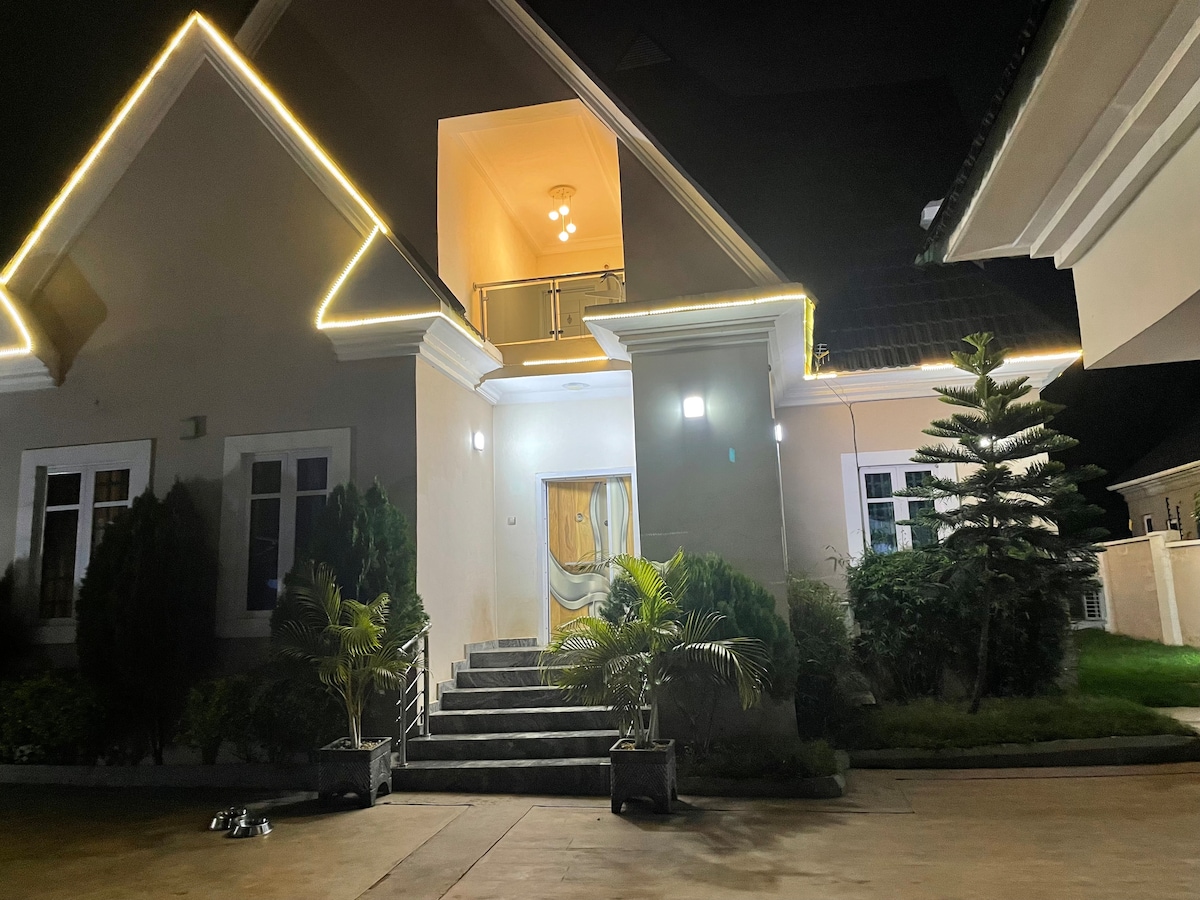 The exterior of a tastefully furnished country home is illuminated by soft outdoor lighting. The entrance features stone steps leading to a welcoming door, framed by well-maintained greenery and potted plants. A balcony is visible on the upper level, adding to the home's charm.