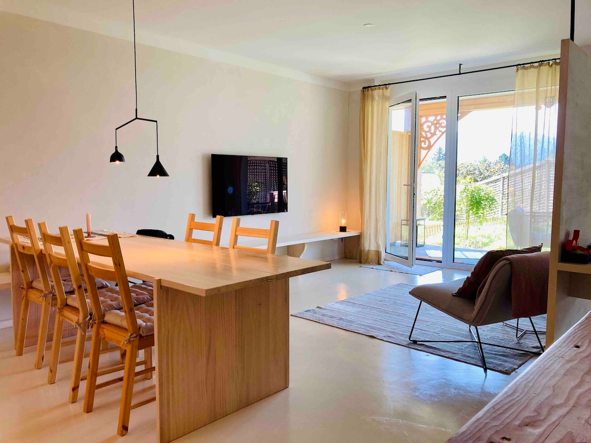 The spacious living area features a large wooden dining table surrounded by six simple chairs. Large glass doors lead to a bright outdoor space, while a flat-screen TV is mounted on the wall. Soft furnishings and gentle lighting add comfort to the room.