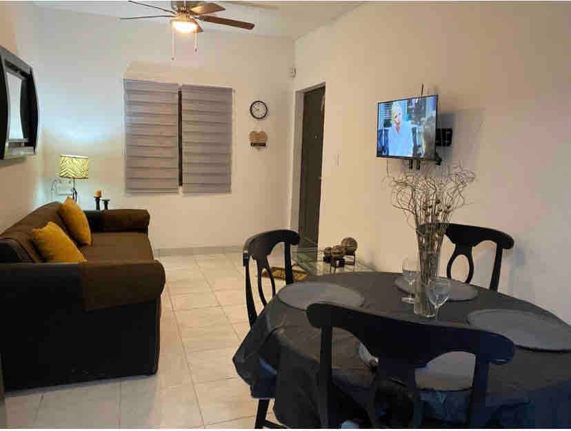 A living area features a dark couch with bright yellow cushions, a dining table set for two, and a television mounted on the wall. Natural light filters through the window with shades, creating a soft ambiance. A decorative plant adds a subtle touch.
