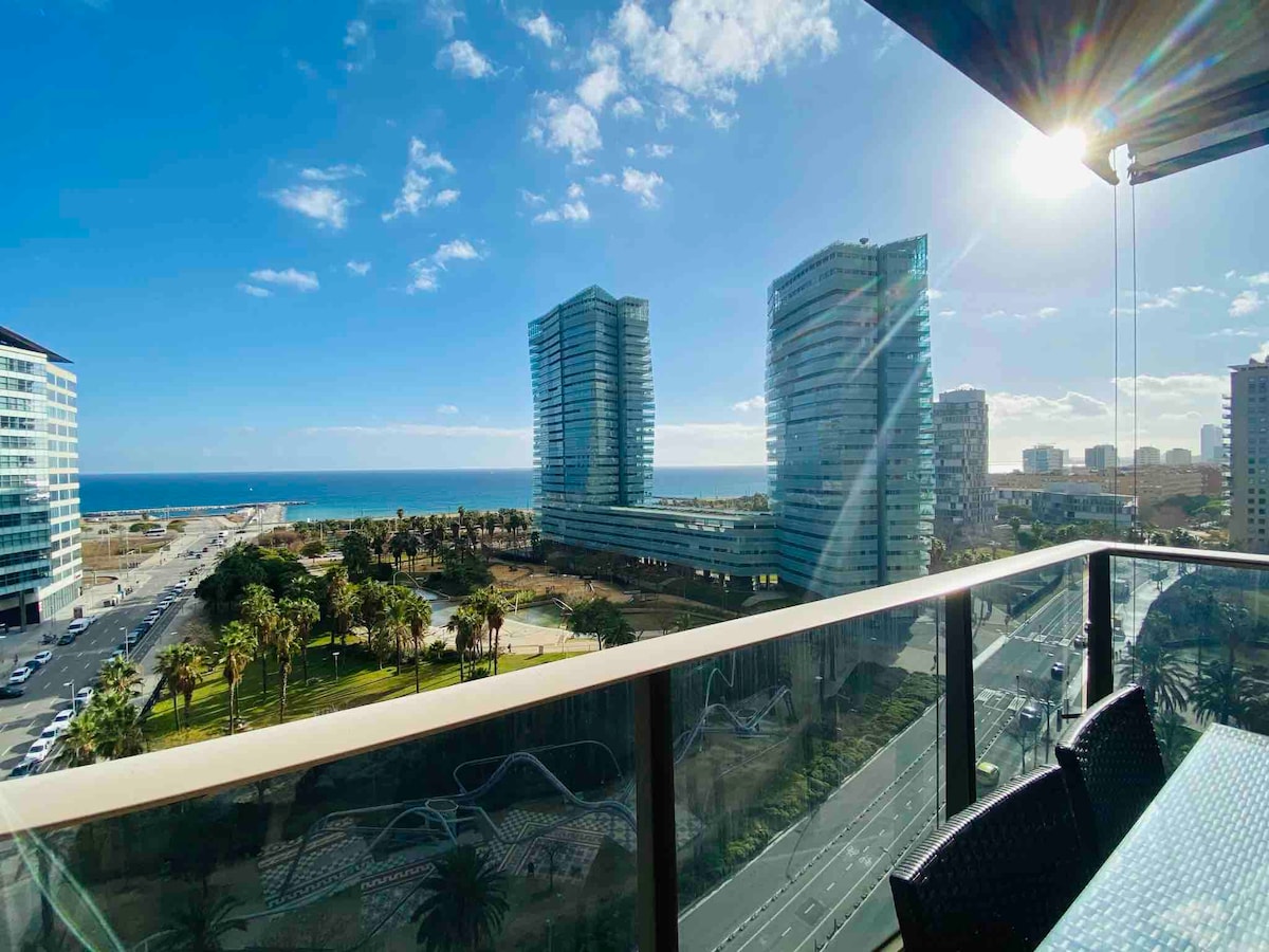 A spacious balcony offers views of the sea and city skyline, framed by modern buildings. Palm trees and greenery are visible in the foreground, along with a clear blue sky scattered with clouds. A dining set is placed on the balcony, inviting relaxation.