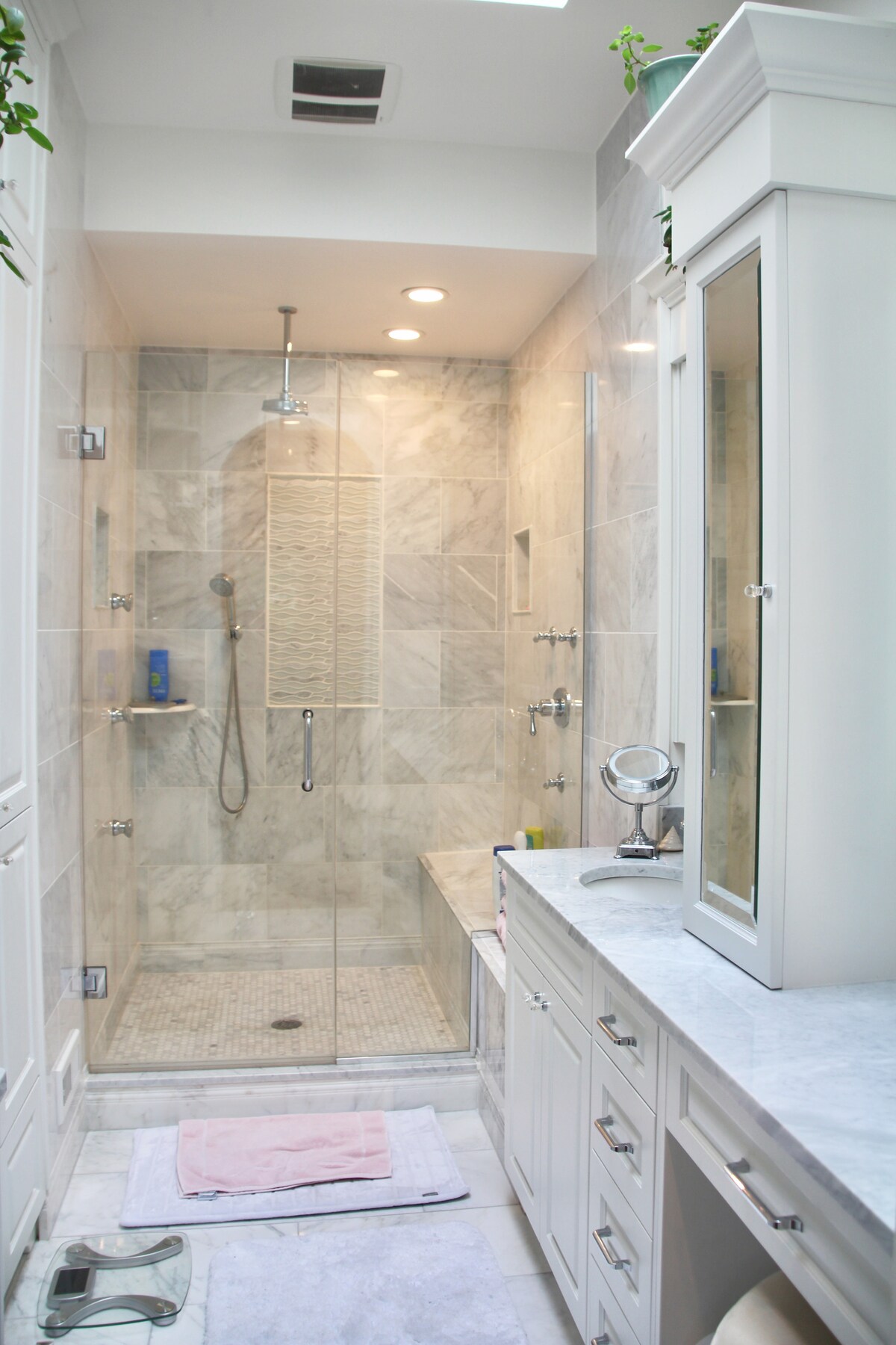 A modern bathroom features a spacious walk-in shower with glass doors and marble walls, providing a clean and bright atmosphere. A double vanity with cabinetry and a large mirror is complemented by soft lighting. A plush bath mat lies on the floor near the vanity.