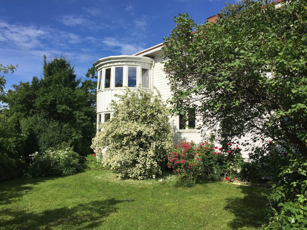 A charming exterior view of a white house, featuring a rounded bay window. Surrounding greenery includes various shrubs and flowering plants, with a well-maintained lawn. Clear blue skies can be seen above, enhancing the serene, natural setting.