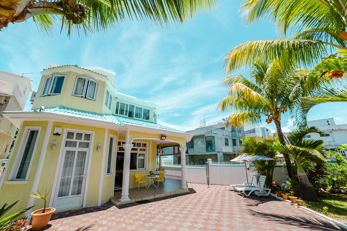The exterior of a two-story villa is showcased, featuring a pale yellow façade and turquoise roof. Palm trees surround the property, providing shade to a patio area with seating. A paved pathway leads towards the entrance, with glimpses of the beach visible in the distance.
