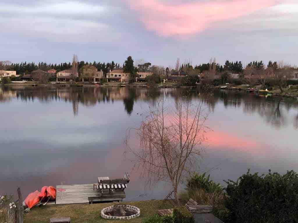 A calm lake reflects the soft hues of a pastel sky at dusk. A small dock with seating faces the water, while a fire pit and a kayak are nearby, allowing for peaceful outdoor gatherings. Surrounding greenery enhances the natural setting.