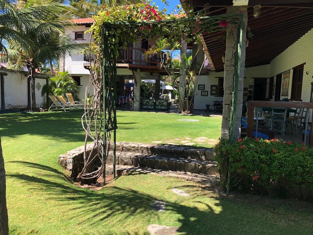 A pathway leads through well-maintained grass, bordered by vibrant tropical plants. A low stone wall is visible beside the steps, creating a transition toward the outdoor area. In the background, seating areas and the two-story structure can be seen, surrounded by palm trees.