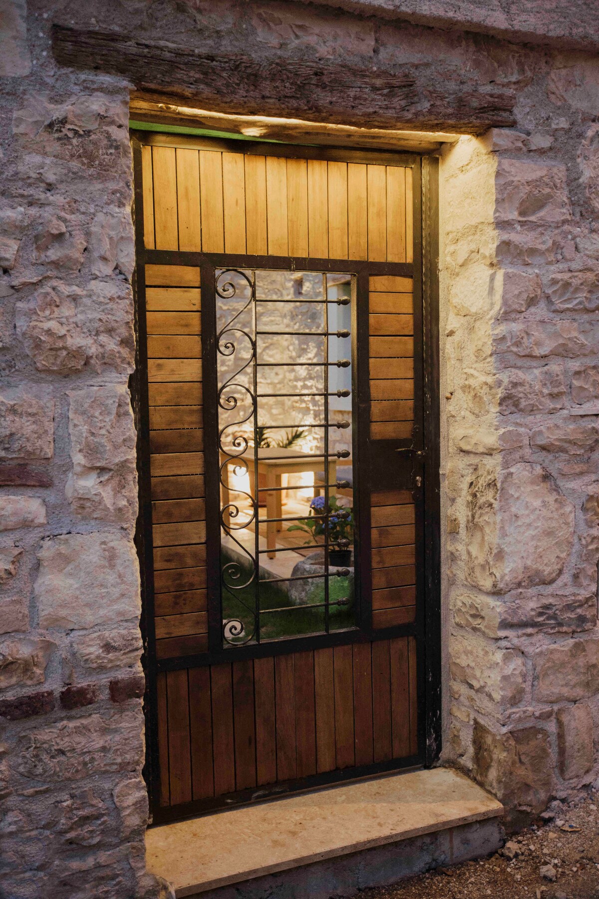 A rustic entrance door made of wood and metal is framed by a stone wall, featuring intricate wrought iron detailing. Soft lighting illuminates the entryway, hinting at the inviting space beyond, with glimpses of greenery and warm decor visible through the open door.