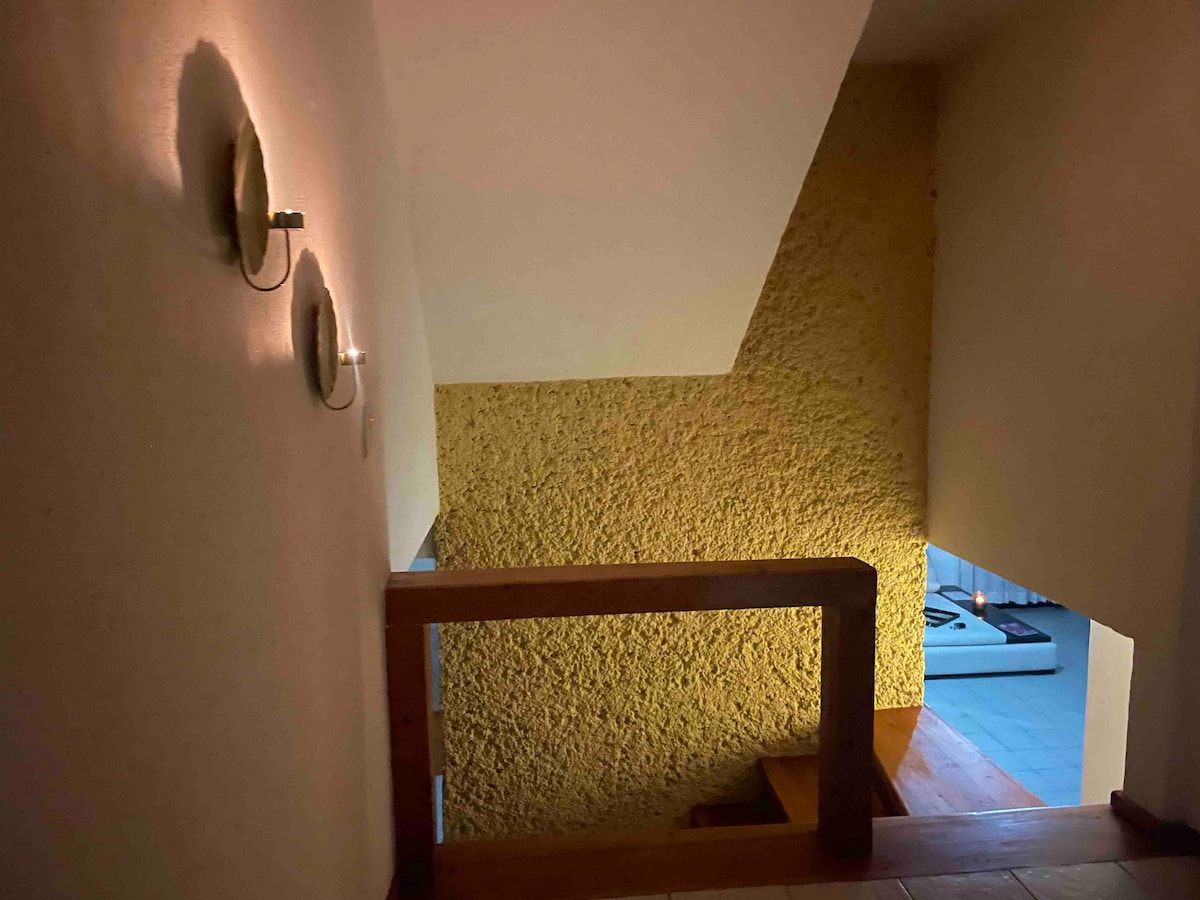 A cozy staircase is seen, featuring textured yellow walls and minimalist sconces providing soft lighting. A wooden railing offers safety and defines the space, while the lower area opens into a softly lit room, creating a warm and inviting passage.