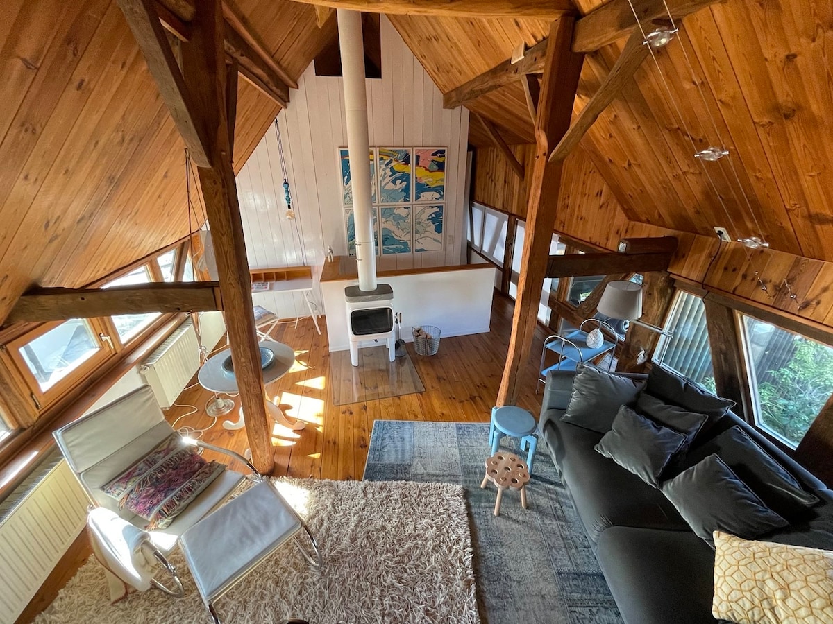 The open living area features exposed wooden beams and large windows, allowing natural light to fill the space. A cozy seating arrangement includes a dark sofa and accent chair. A small table and wood stove are included, enhancing the room’s charm with a warm ambiance.