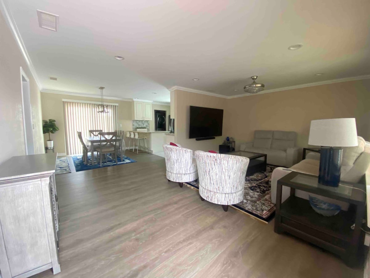 The living area is equipped with a large flat-screen TV mounted on the wall, surrounded by comfortable seating. A dining table for six is visible in the background, adjacent to the kitchen. Ample natural light enters through sliding glass doors leading to an outdoor space.