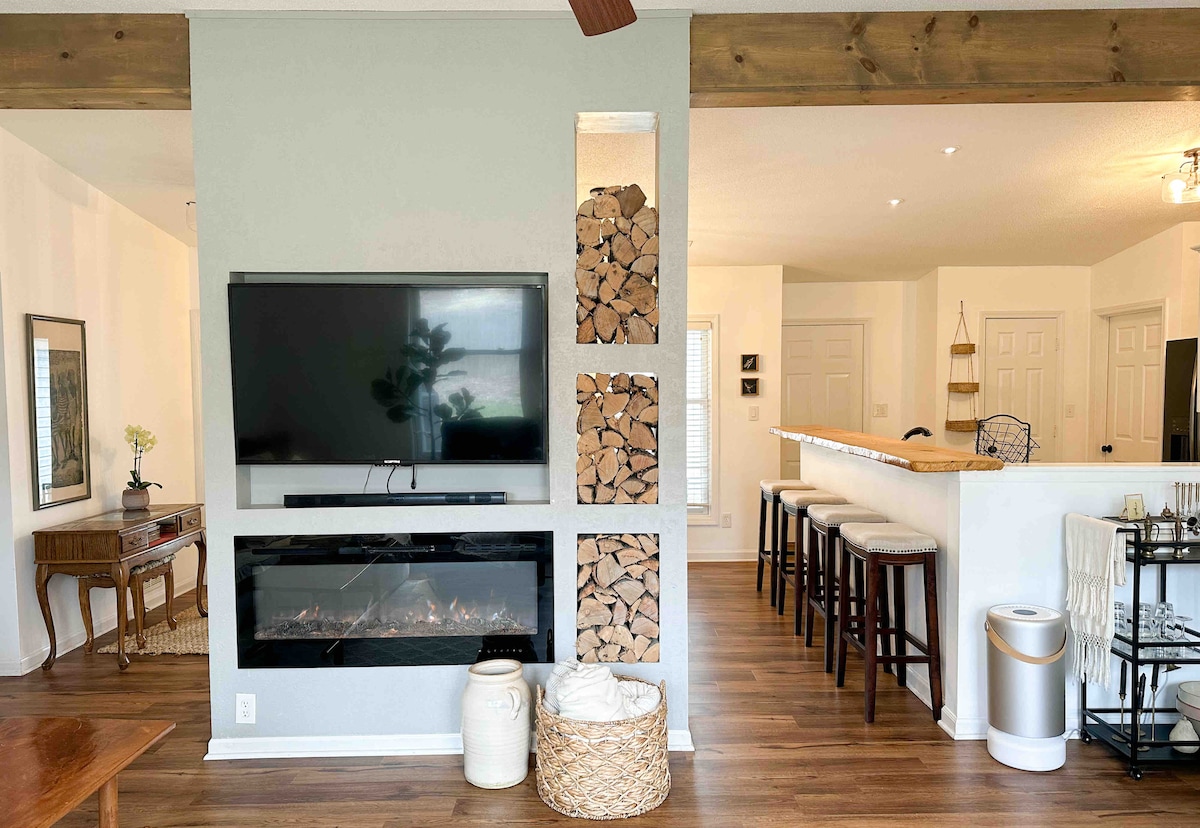 A modern living area features a wall-mounted television above a sleek electric fireplace. Stacked firewood is neatly displayed in a recessed area. The room flows into a bright kitchen, with a breakfast bar highlighted by high stools. Natural light floods the space from nearby windows.