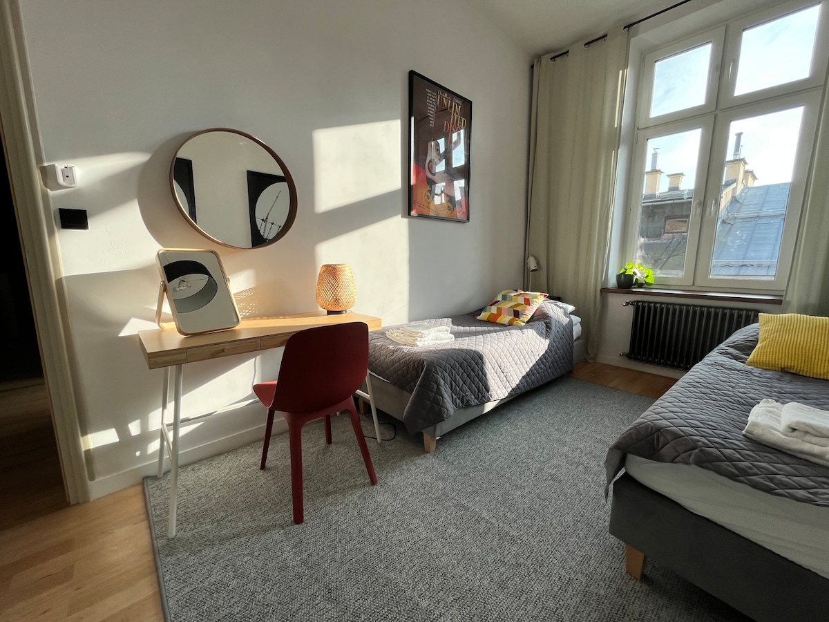 The bedroom features two single beds, each made with gray blankets. A desk is positioned beside a circular mirror, accented by a red chair. Natural light filters through the large windows, illuminating the room and highlighting a decorative wall art piece.