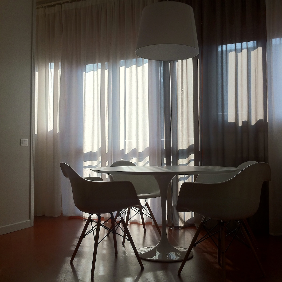 A dining area is highlighted by a circular white table surrounded by four modern chairs. Sheer curtains diffuse natural light, creating a soft ambiance. Large windows offer views of the outside while maintaining a sense of privacy.