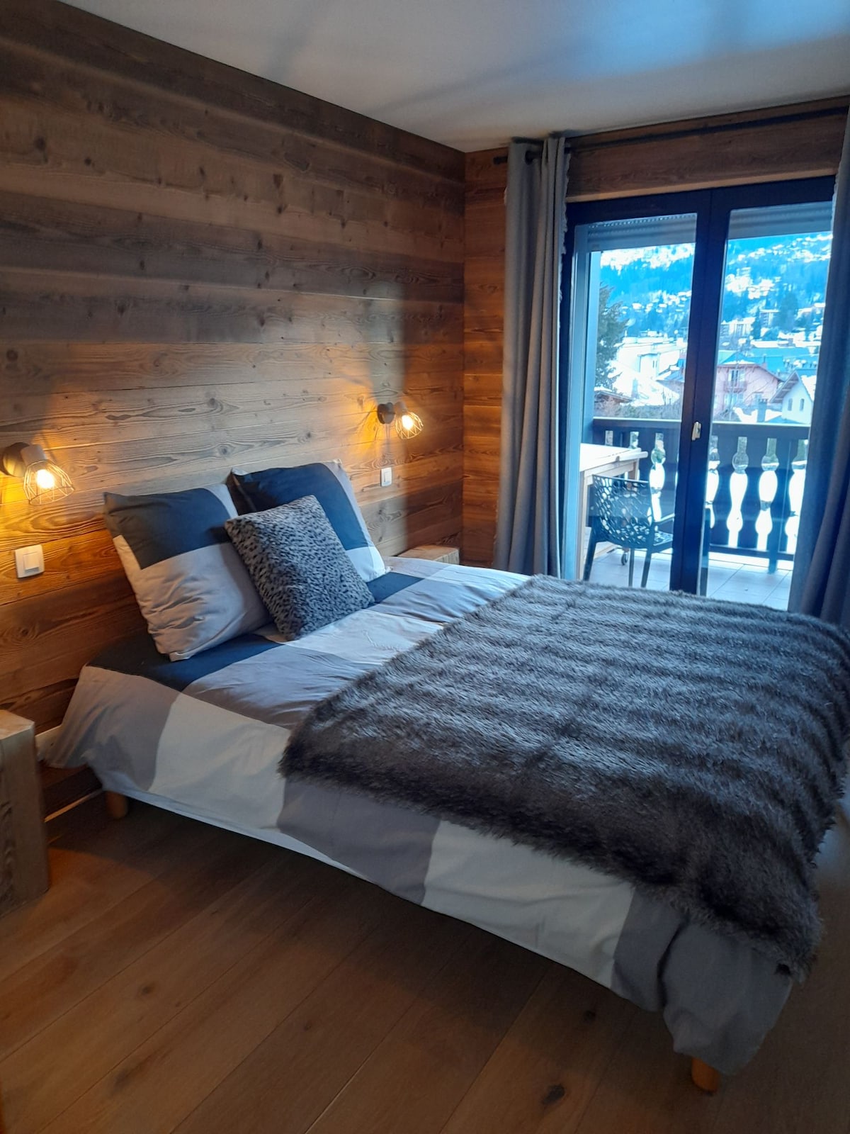 A cozy bedroom features wooden accent walls and a spacious bed layered with soft bedding and a textured throw. A sliding glass door leads to a balcony overlooking the serene outdoor landscape, while bedside lamps provide warm, ambient lighting.