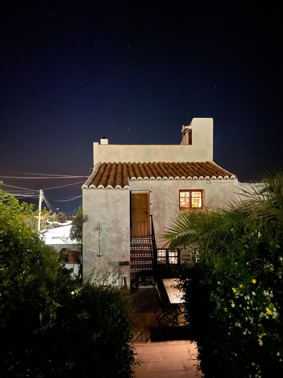 A quaint cottage exterior is illuminated at night, revealing a terracotta tiled roof and a wooden door. A staircase leads to the upper level, while surrounding greenery creates a serene, private atmosphere. Stars can be seen in the night sky above the structure.