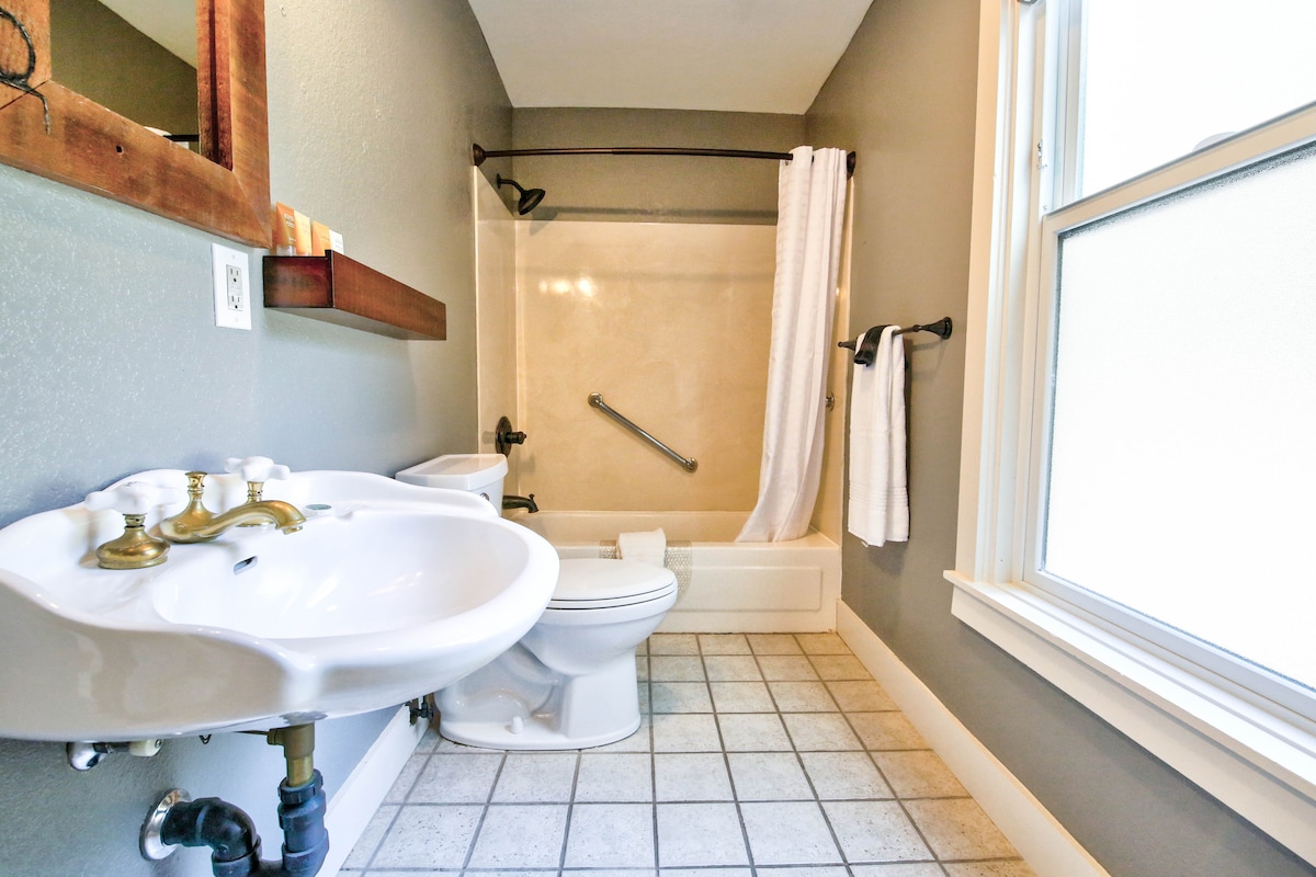 The bathroom features a classic design with a dual sink setup and a toilet. A combination bathtub and shower is equipped with grab bars and a shower curtain. A window allows natural light. Towel racks are mounted beside the tub, and a wooden shelf is positioned above the sinks.