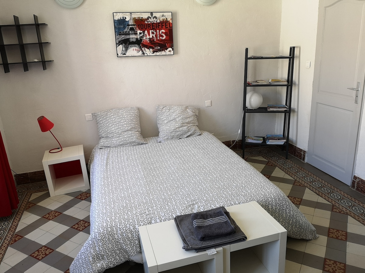 A spacious sleeping area features a light-colored patterned duvet covering a double bed with two matching pillows. A red bedside lamp and a white shelf with a round lamp stand are positioned nearby. Decorative artwork depicting Paris is mounted on the wall.
