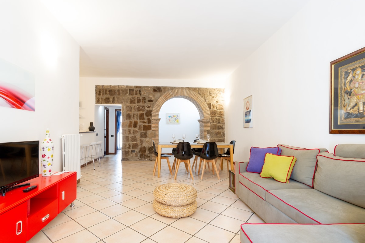 A spacious living area is highlighted by a neutral color palette and natural stone wall, creating a warm ambiance. A comfortable sofa adorned with colorful cushions faces a red cabinet and a television. A dining table with black chairs is set for casual gatherings.