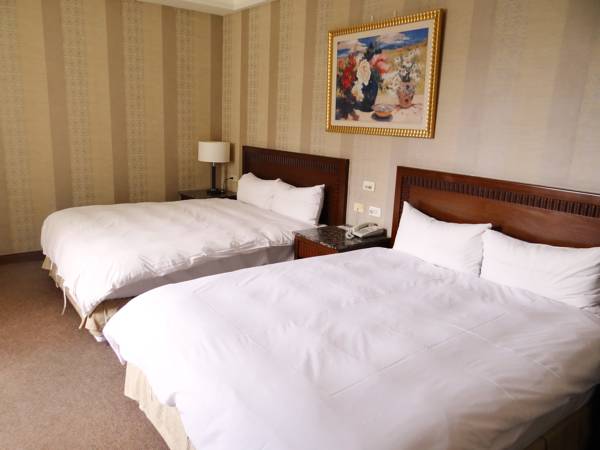 The room features two spacious beds with white comforters, positioned beside a wooden headboard. A bedside table with a lamp and phone is visible between the beds. Artwork adorns the wall above, complementing the neutral striped wallpaper.
