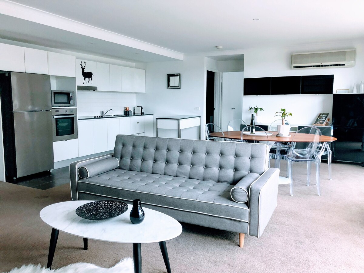 An open-plan living area is presented with a modern design. A comfortable gray sofa is positioned in front of a round marble coffee table, complemented by clear dining chairs around a wooden dining table. The kitchen is equipped with sleek appliances, including an oven and a refrigerator.