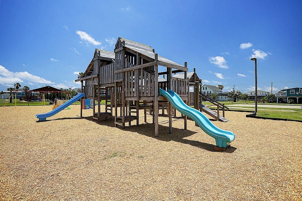 A wooden playground structure is featured, incorporating multiple slides in vibrant blue shades. A sandy surface surrounds the equipment, providing a safe play area. The background shows open grassy spaces under a clear blue sky, enhancing the inviting setting for outdoor activities.