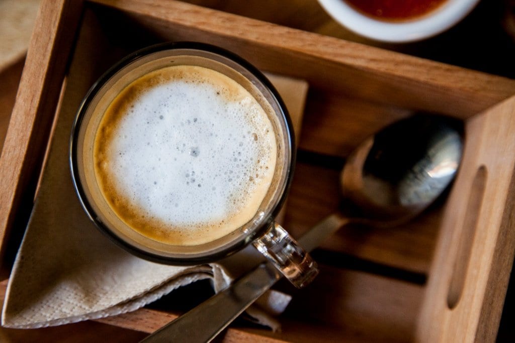 A glass of espresso is presented atop a wooden tray, accompanied by a folded napkin. A stainless steel spoon mirrors the glass's sleek design, enhancing the visual simplicity of the arrangement. Soft foam crowns the rich beverage, inviting attention.
