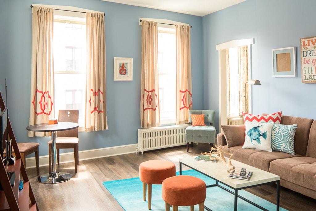 A bright living room features light blue walls and natural light flooding through large windows. A cozy sofa is accented by colorful cushions. A small round dining table with stylish chairs sits next to the sofa, complemented by two orange ottomans and a contemporary wooden shelf.