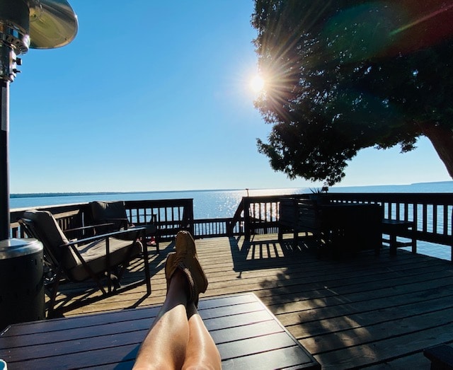 A sunlit deck features comfortable seating arrangements oriented towards the tranquil water. A glimpse of the sun is visible on the horizon, casting serene reflections on the surface. Expansive views allow for enjoyment of the calming surroundings while relaxing outdoors.