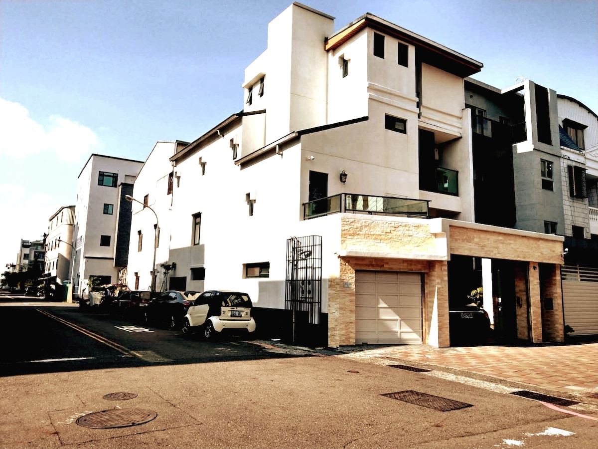 The exterior of a modern building features a clean design with multiple levels and large windows. A stone entrance leads to an attached garage, while parked cars line the street. The clear sky casts light on the façade, giving it a bright appearance.
