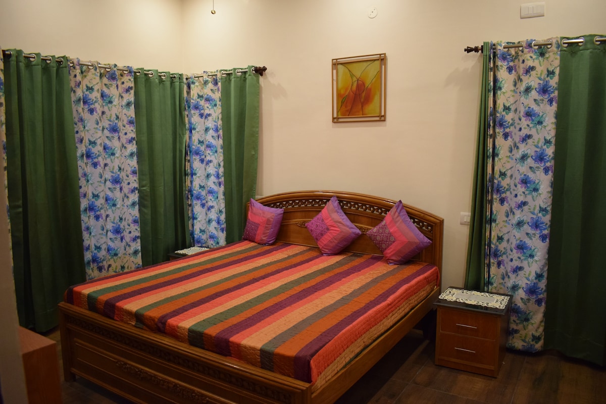 A spacious sleeping area features a wooden bed adorned with a striped mattress and decorative pillows. Vibrant, floral-patterned curtains frame the window, complemented by green drapes, creating a colorful and inviting ambience.