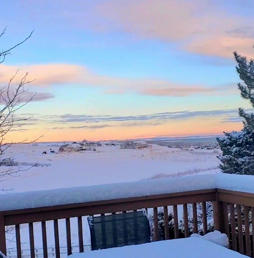 A serene winter landscape is visible from the deck, showcasing a snow-covered expanse under a pastel sky at sunrise. Soft hues of pink and orange blend with the fading blue, creating a peaceful atmosphere in this tranquil outdoor setting.