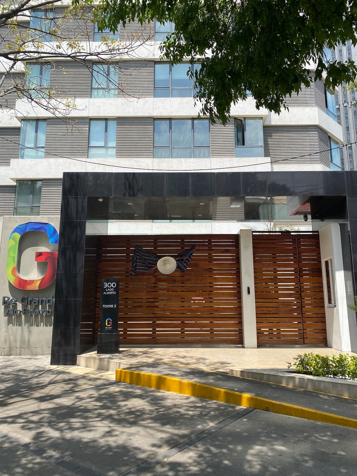The entrance features a modern facade with a large wooden gate and glass elements. The name 'G' is displayed prominently beside the entrance. A well-maintained sidewalk leads up to the entrance, surrounded by landscaped greenery.