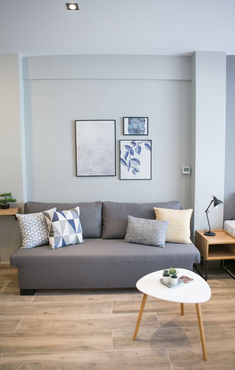 A modern living room is displayed, featuring a gray couch adorned with patterned cushions. A small round table is positioned in front, holding a small plant. The wall is decorated with three framed artworks, adding a touch of creativity to the space.