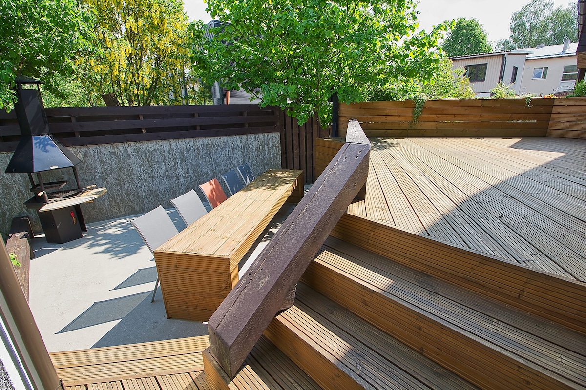 A wooden terrace is presented, featuring a series of built-in seating areas and a charcoal barbeque grill. Steps lead down to a lower level, surrounded by trees and greenery, creating a calm outdoor setting for gatherings.