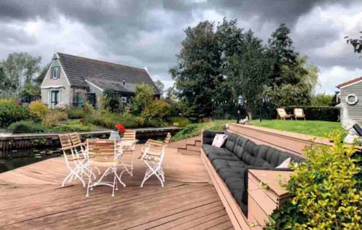 An outdoor decking area features a spacious lounge couch surrounded by greenery, creating a serene setting. A dining table with chairs is set for gatherings. The scene captures the tranquility of the nearby water, with the house visible in the background.