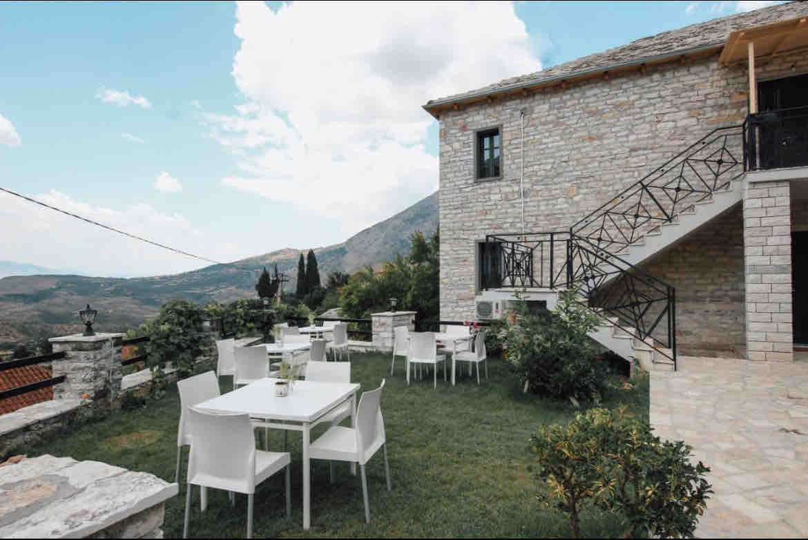 An outdoor terrace features several white chairs and tables arranged on a well-maintained lawn. Stone walls surround the area, and the scenic backdrop includes rolling hills and a cloudy sky, adding to the inviting atmosphere for guests.