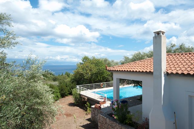 A villa is set amidst lush greenery, with a private swimming pool visible in the backyard. The clear blue sky is reflected in the water, while the charming red-tiled roof and white exterior of the villa contrast with the surrounding landscape.