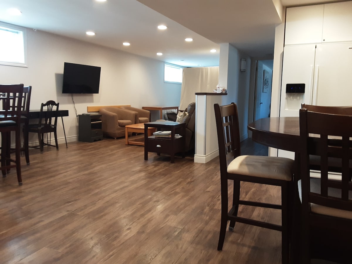 An open-concept living space is shown, featuring a large flat-screen television mounted on the wall and comfortable seating arranged around a coffee table. Dining areas with high chairs are visible, along with a well-equipped kitchen in the background. Natural light enters through several windows.