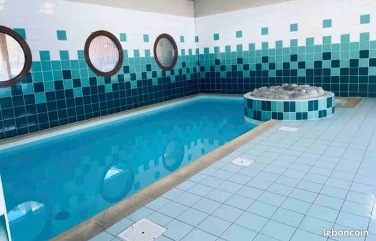 An indoor pool area features a sleek tiled design in shades of blue. Round windows allow natural light, while a seating area with smooth stones is positioned alongside the pool, creating a serene environment for relaxation.