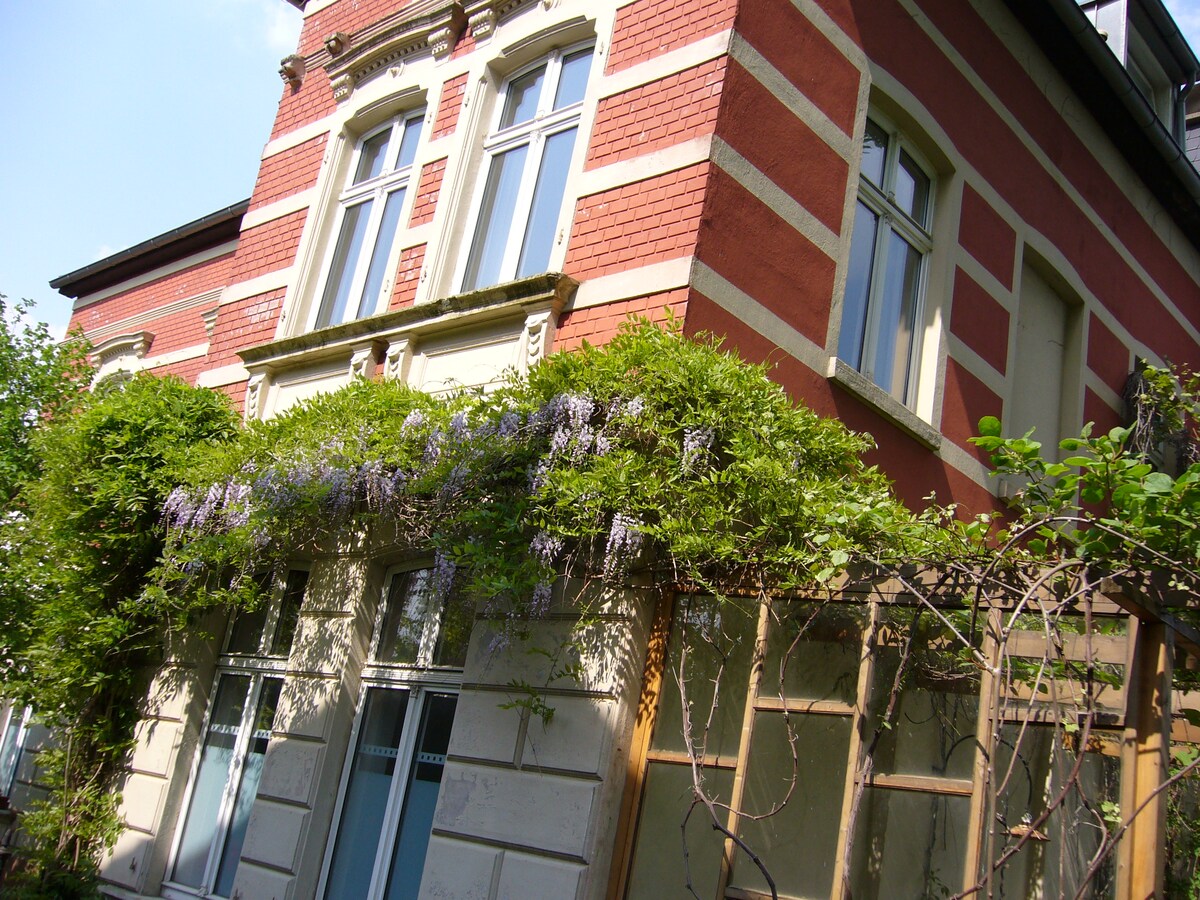 A historic building is featured, showcasing a mix of red brick and white stone. Elegant windows adorn the façade, framed by lush greenery and blooming wisteria, enhancing the building's classic charm.
