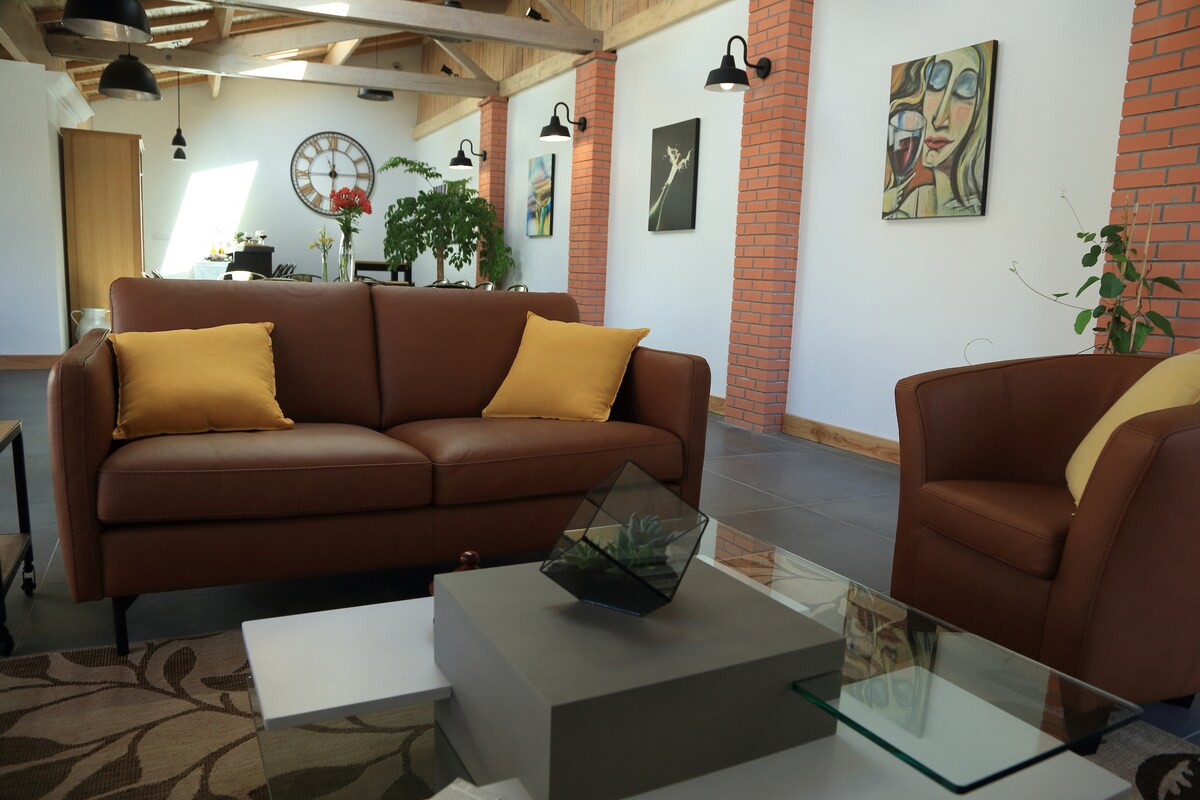 A comfortable seating area is highlighted by a brown leather sofa adorned with yellow cushions. A glass coffee table sits in front, complemented by a small decorative item. The walls feature a warm brick design and are illuminated by stylish wall lamps.