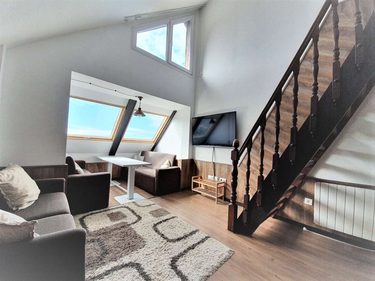A bright living area is shown, featuring modern furniture, including a light table surrounded by two cushioned seating arrangements. Large, angled windows allow natural light to fill the space, with a staircase leading to an upper level visible in the background.