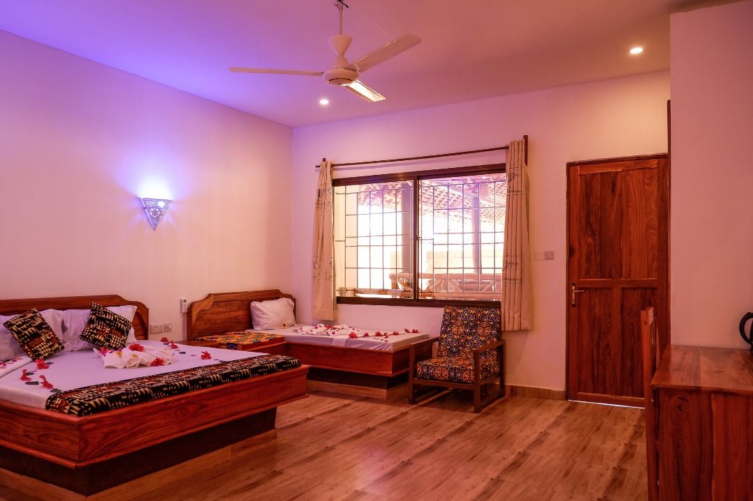 A spacious room is equipped with a king size bed and a single bed, adorned with decorative petals. A wooden wardrobe and a writing desk with a chair are present. Natural light fills the room through a large window, complemented by warm wall lighting.