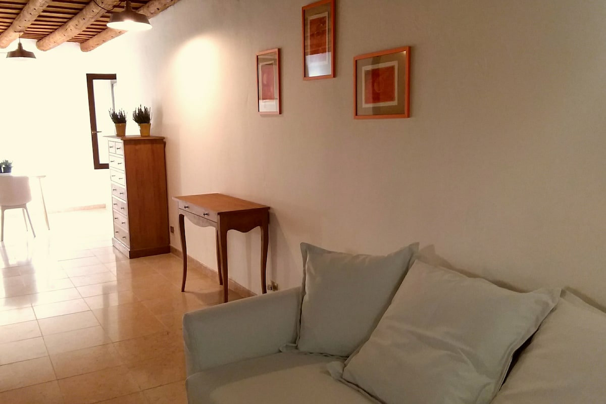 A bright living area showcases a comfortable sofa with soft cushions, complemented by a wooden side table. The walls are adorned with simple framed art, while natural light fills the space through a window, enhancing the airy atmosphere.