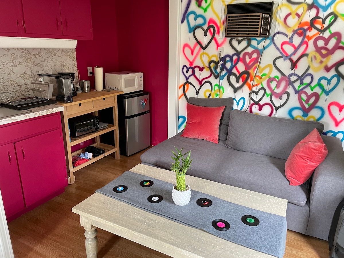 A vibrant guest studio features pink accent walls adorned with colorful heart graffiti. A compact kitchenette with essential appliances is visible. A comfortable gray sofa is placed beside a light wooden table showcasing a potted plant and a decorative table runner inspired by vinyl records.