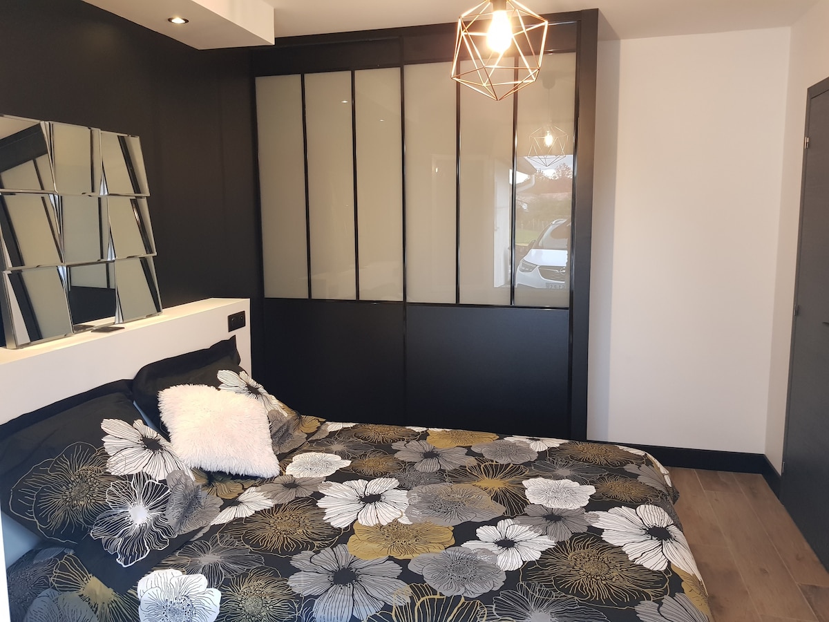 The bedroom features a spacious bed with a floral-patterned duvet cover, complemented by a soft pillow. A geometric light fixture hangs above, and mirrored panels add a modern touch. The dark accent wall provides contrast to the lighter tones of the bedding.